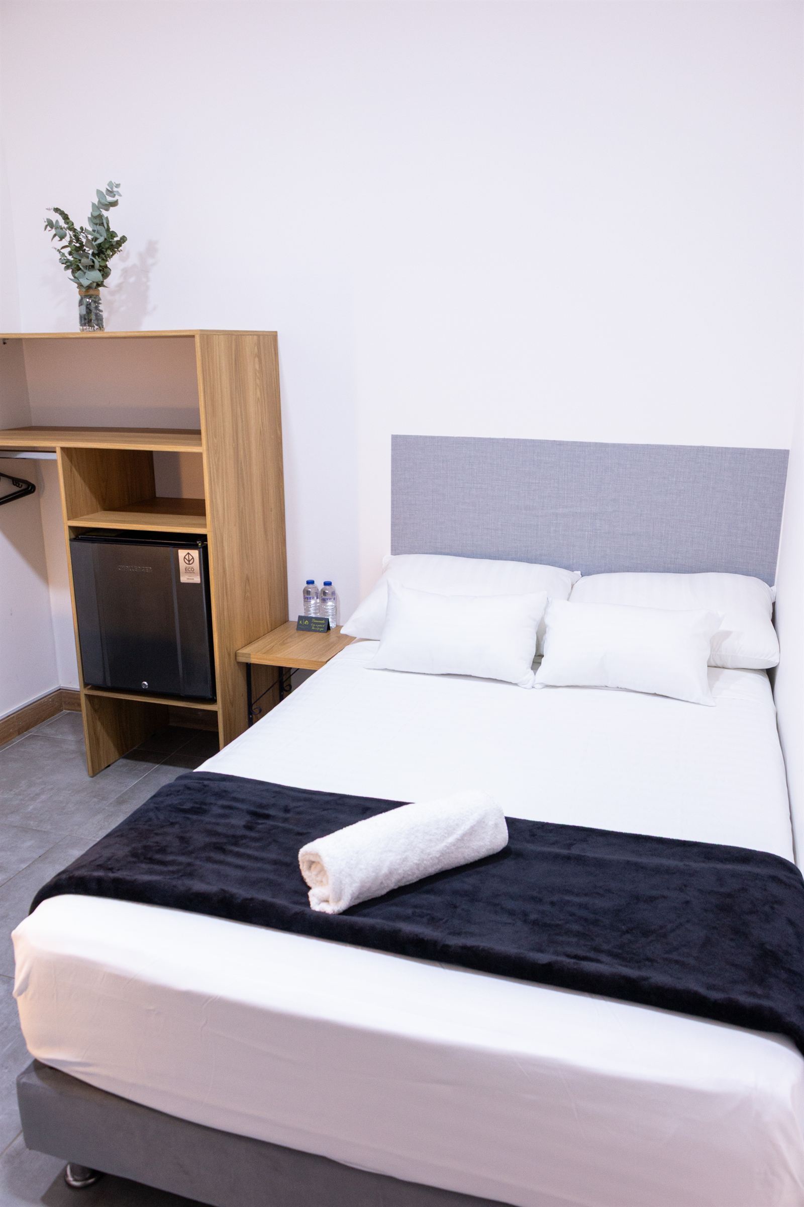 Cama doble con cabecero gris, estante de madera con mini nevera integrada y planta decorativa — Doble Superior Internas